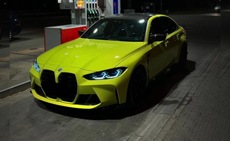 Żółte BMW M3 Competition z włączonymi światłami na stacji benzynowej nocą