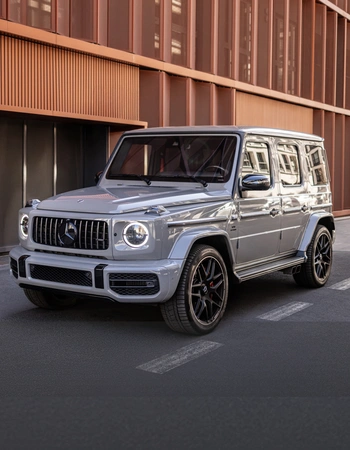 Mercedes G63 AMG in front of modern urban architecture, front view with daytime running lights on.