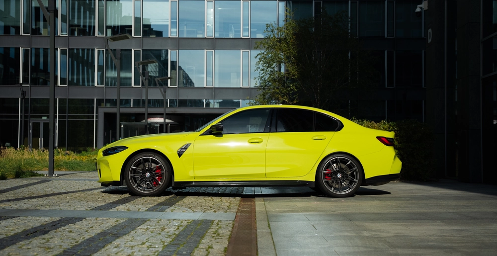 Jaskrawożółte BMW M3 Competition xDrive z profilu, na tle nowoczesnego biurowca.
