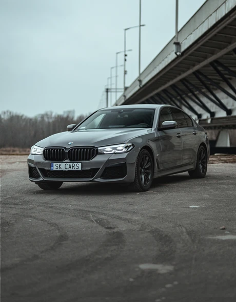 Szary BMW 5 Series na tle miejskiego krajobrazu pod mostem. Styl i moc dostępne do wynajmu w SK CARS, idealne na podróże miejskie.