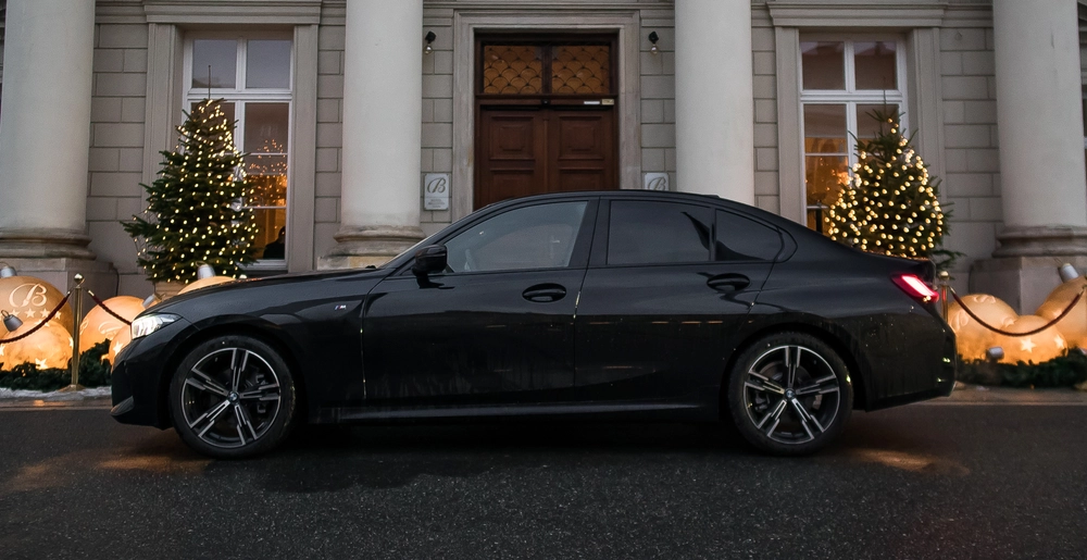 BMW 3 Series z eleganckim wyglądem przed stylową fasadą, podkreśla świąteczny klimat dzięki światłom i choinkom. Wynajem dostępny na stronie SK Cars.