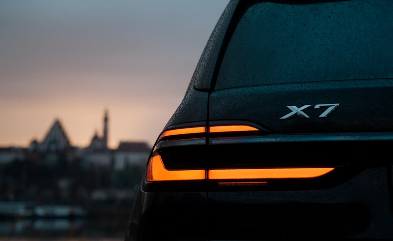 Tylne światła BMW X7 xDrive 40 w deszczu – luksusowy SUV nocą z panoramą Warszawy w tle. Wynajem samochodów premium – SK CARS.