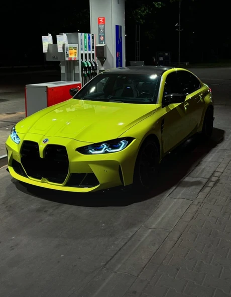 Żółte BMW M3 Competition z włączonymi światłami na stacji benzynowej nocą