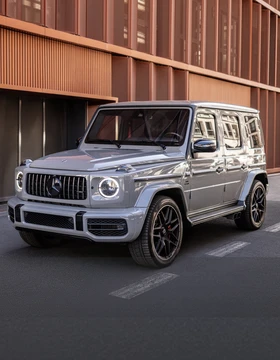 Mercedes G63 AMG na tle nowoczesnej architektury miejskiej, widok z przodu z włączonymi światłami do jazdy dziennej.