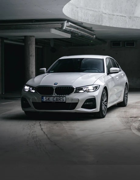 Przednia część białego BMW 3 Series na stylowym parkingu podziemnym. Elegancja i moc dla Twojej przyjemności z jazdy.